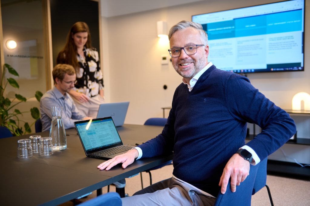 Ein Berufstätiger mit Laptop posiert für ein Foto und zwei weitere Berufstätige schauen auf ihren Computer